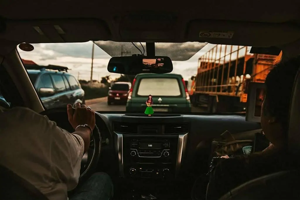 A dashboard hula dancer seen dead-center in a car with a driver and passenger on both sides; it appears that the car is driving through traffic.