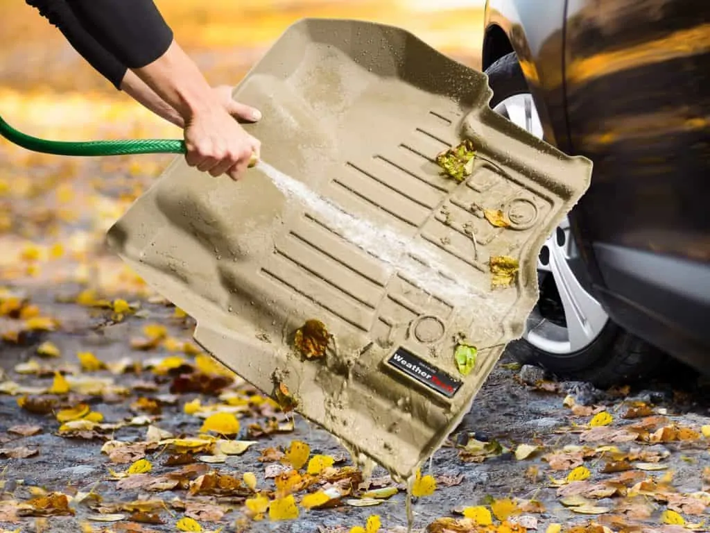 A product image for an easy-clean floor mat, featuring an action shot of a person spraying a hose on the product to wash leaves and dirt onto the ground. The vehicle is seen partially on the right side to demonstrate the purpose of the product. 